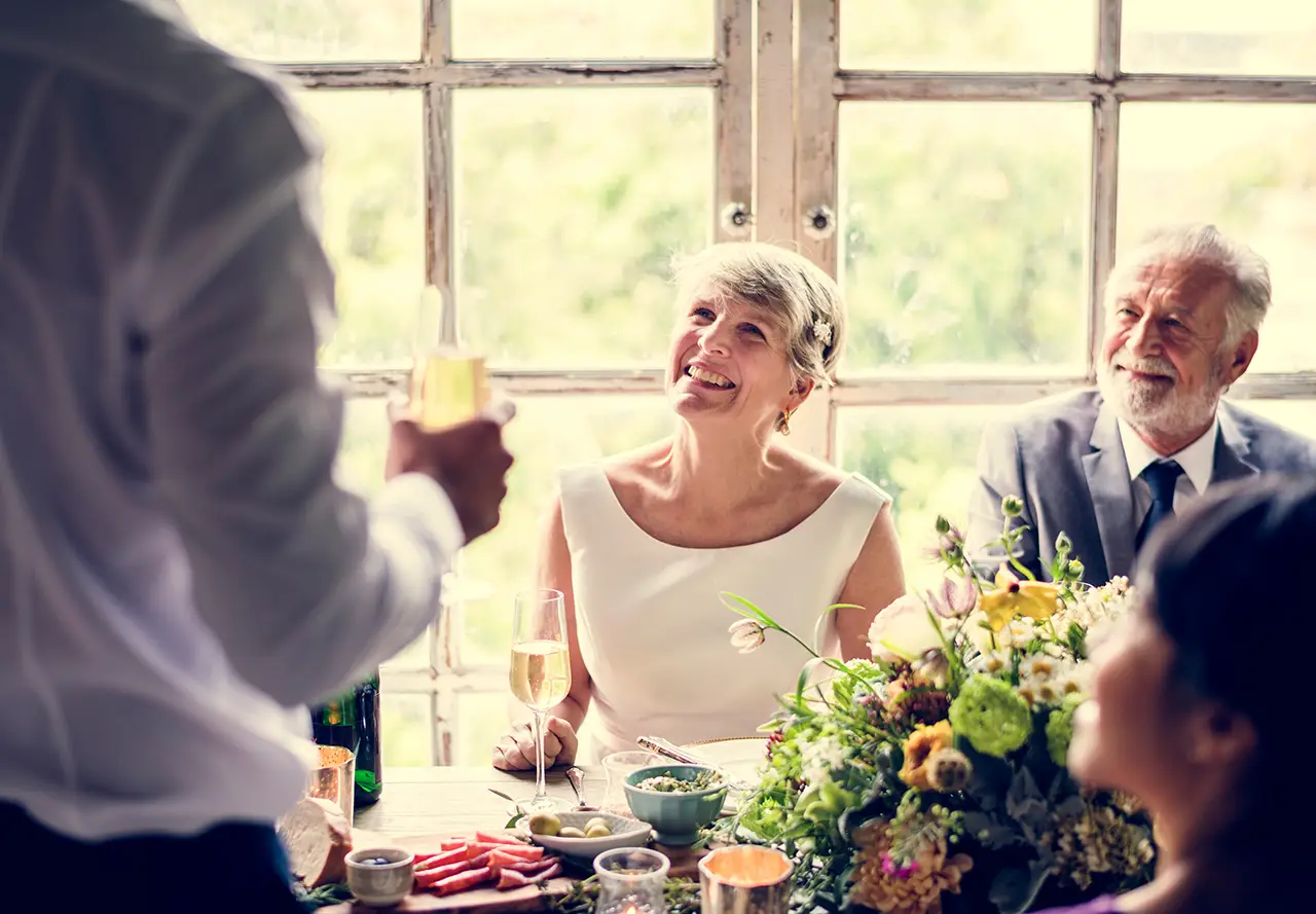 een ouder bruidspaar zit lachend aan tafel terwijl iemand niet zichtbaar een speech geeft aan het bruidspaar