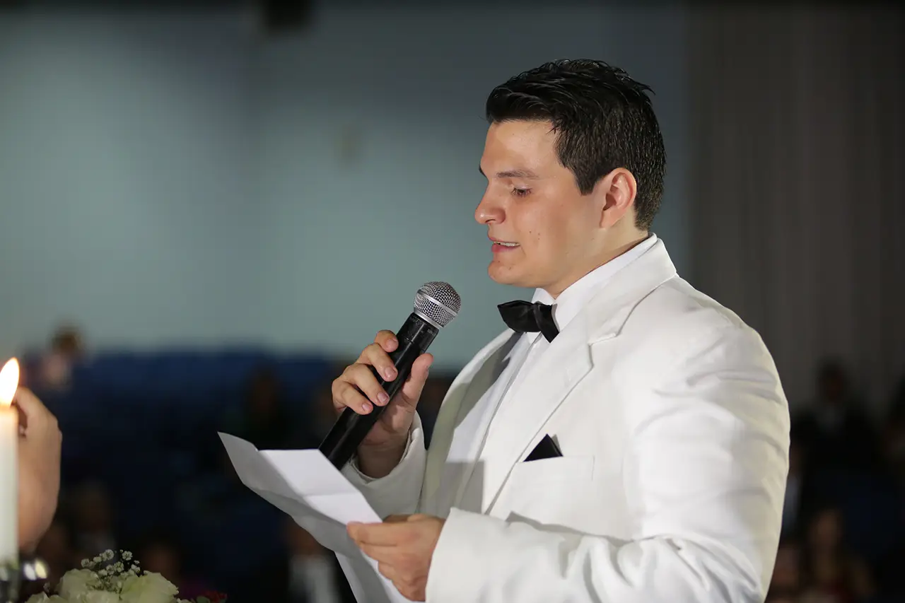 een man in een wit trouwpak geeft een speech vanaf een briefje met een microfoon in zijn handen